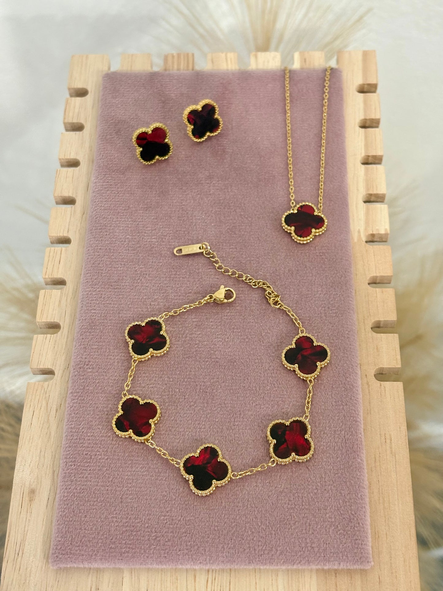 3-piece gold stainless steel jewelry set featuring deep red enamel four-leaf clover necklace, stud earrings, and multi-motif bracelet on a mauve display.