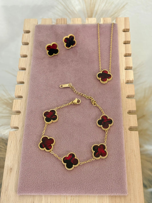 3-piece gold stainless steel jewelry set featuring deep red enamel four-leaf clover necklace, stud earrings, and multi-motif bracelet on a mauve display.
