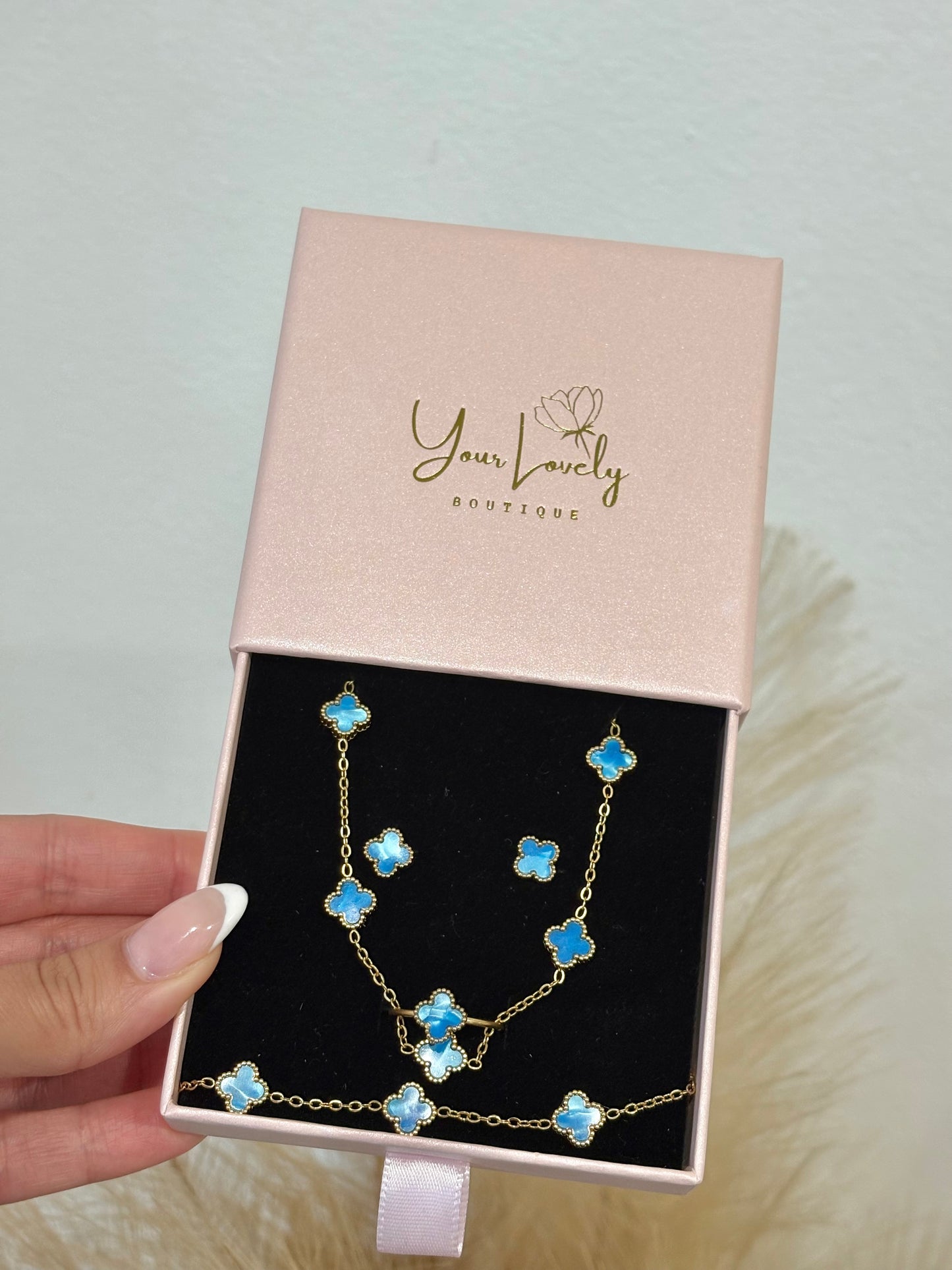 4-piece gold stainless steel jewelry set featuring mini baby blue enamel four-leaf clover 5-motif necklace (16-18 inch), bracelet, stud earrings, and adjustable ring on a purple display.⁠