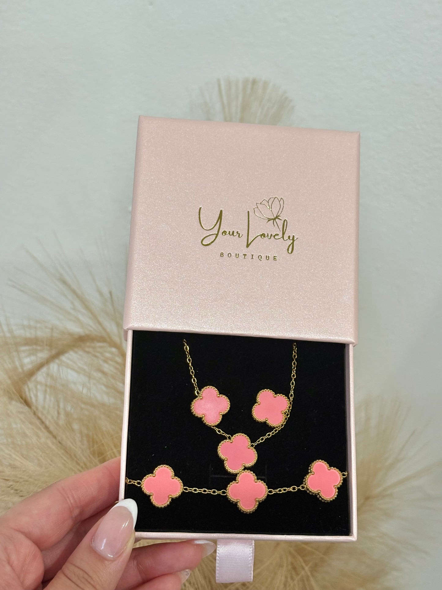 3-piece gold stainless steel jewelry set featuring coral pink enamel four-leaf clover necklace, stud earrings, and five-motif link bracelet on a mauve velvet display.⁠