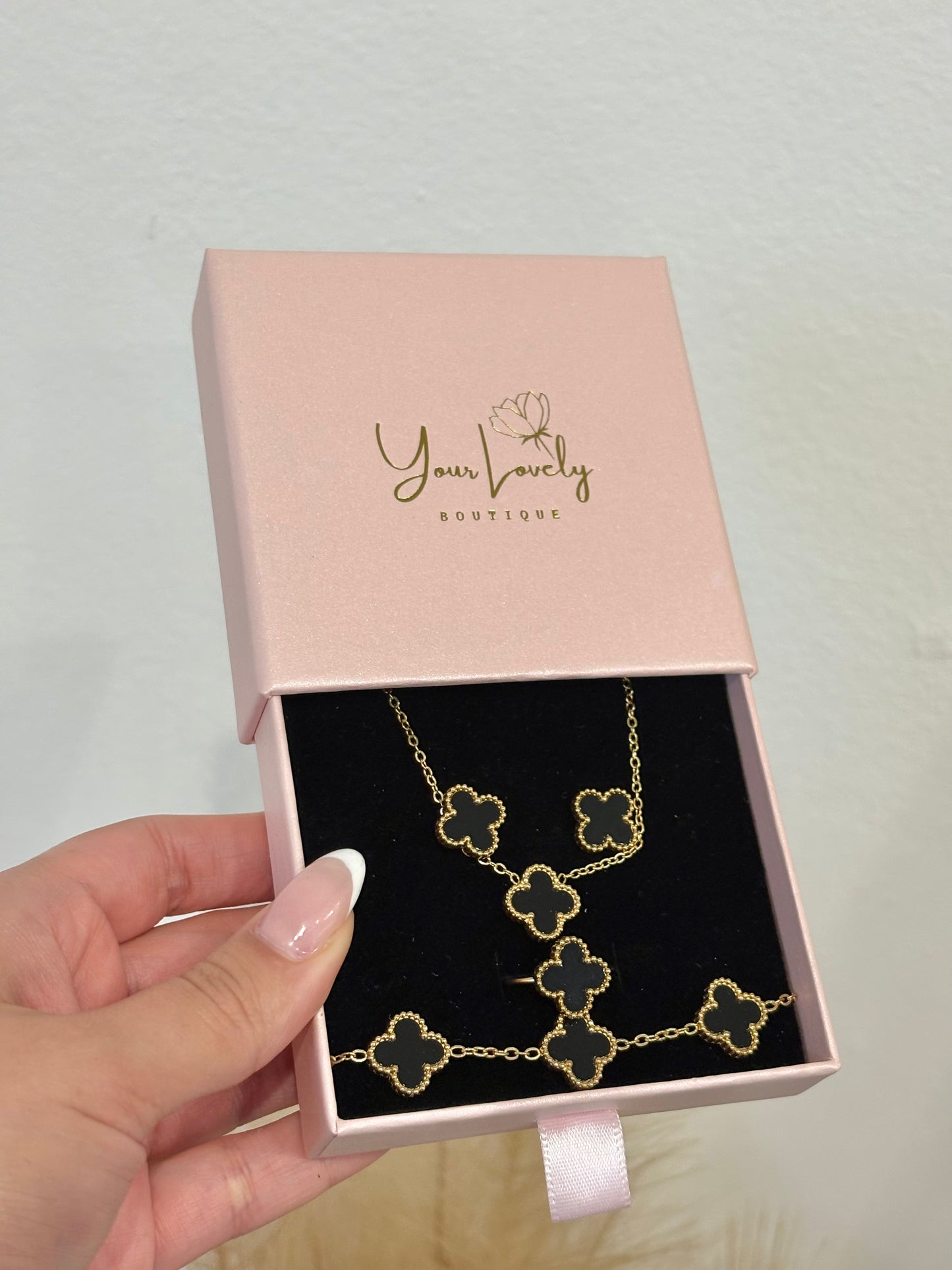 4-piece gold stainless steel jewelry set featuring mini black enamel four-leaf clover necklace, stud earrings, five-motif link bracelet, and adjustable ring on a purple display.⁠