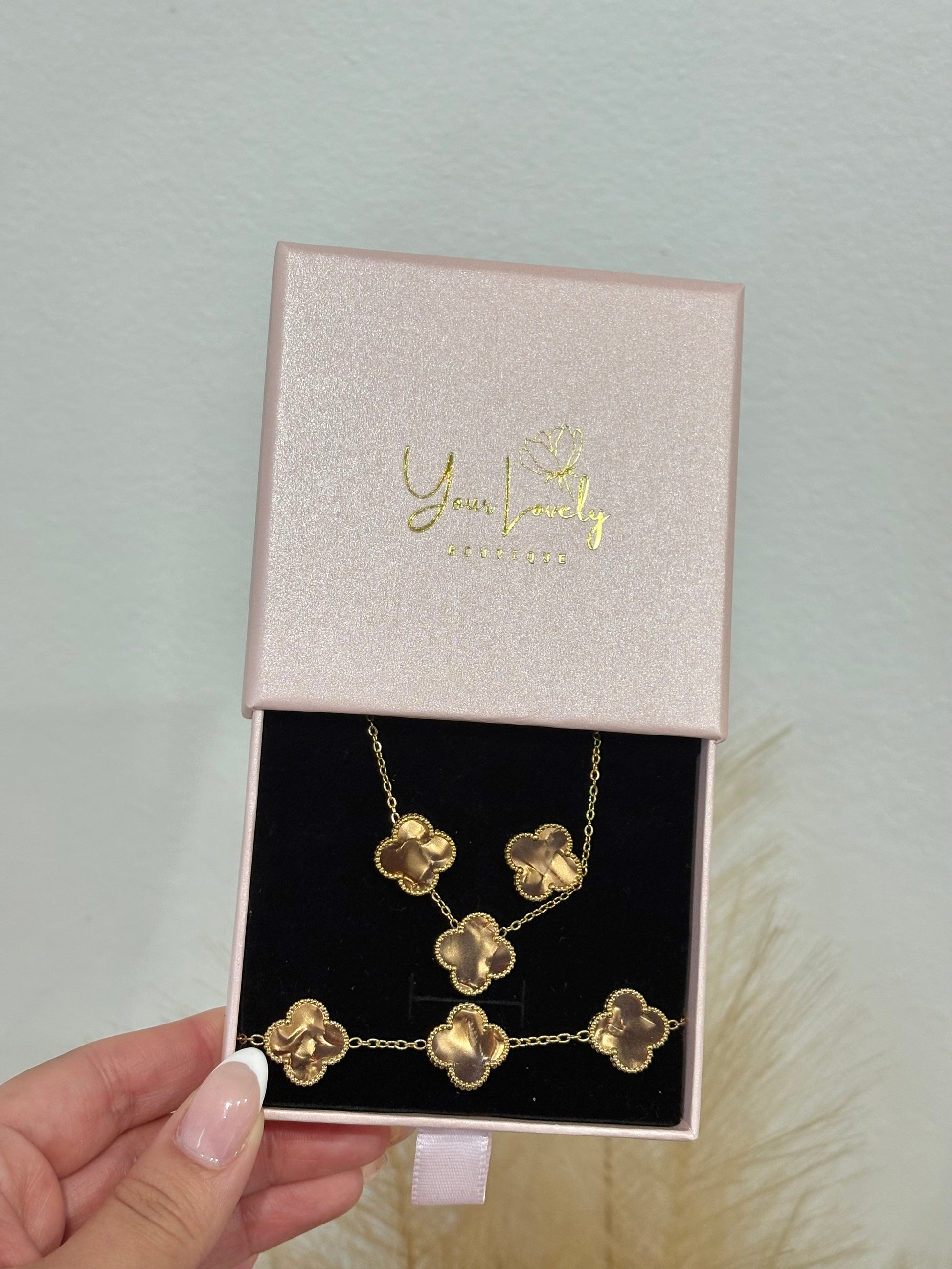 3-piece gold stainless steel jewelry set featuring brown enamel four-leaf clover necklace, stud earrings, and multi-clover bracelet displayed on a purple velvet stand