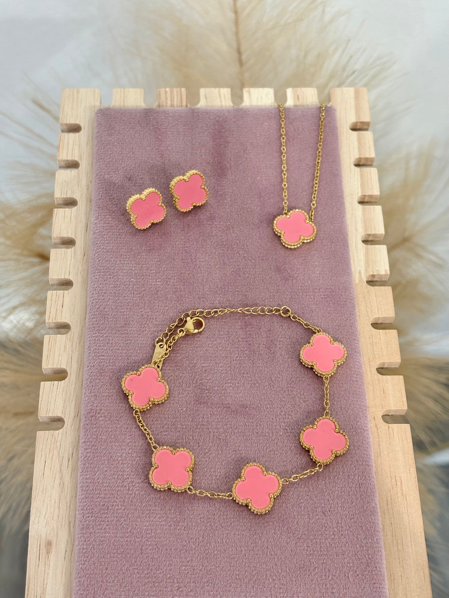 3-piece gold stainless steel jewelry set featuring coral pink enamel four-leaf clover necklace, stud earrings, and five-motif link bracelet on a mauve velvet display.