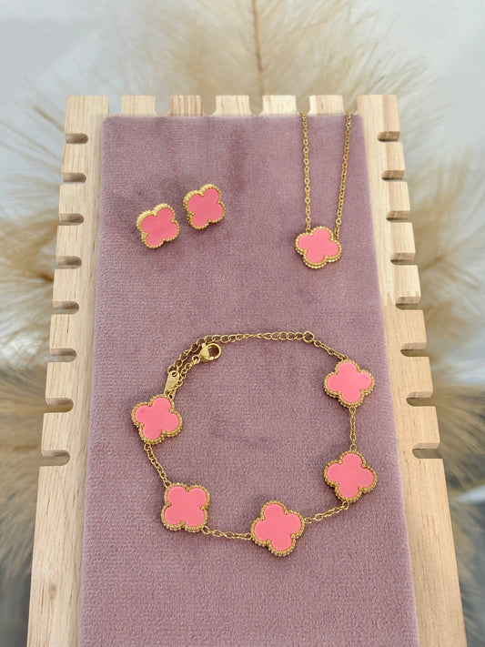 3-piece gold stainless steel jewelry set featuring coral pink enamel four-leaf clover necklace, stud earrings, and five-motif link bracelet on a mauve velvet display.