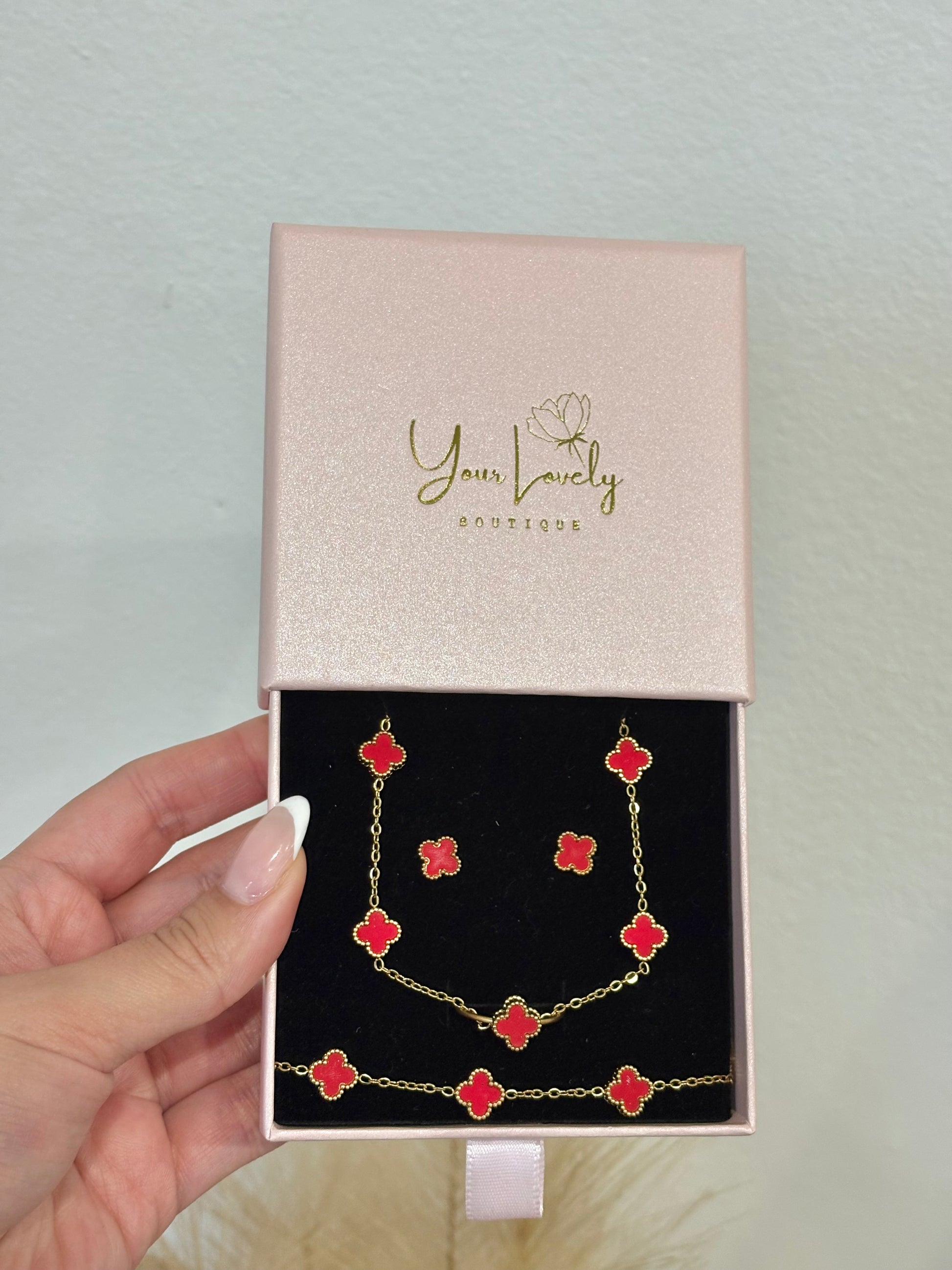 4-piece gold stainless steel jewelry set featuring mini red enamel four-leaf clover 7-motif necklace, 5-motif bracelet, stud earrings, and adjustable ring on a mauve velvet display.⁠