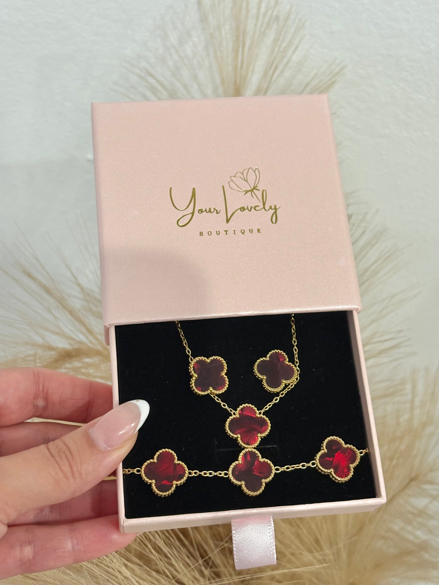 3-piece gold stainless steel jewelry set featuring deep red enamel four-leaf clover necklace, stud earrings, and multi-motif bracelet on a mauve display.