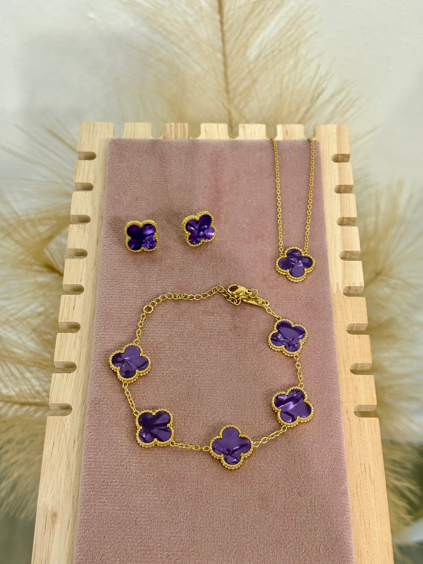 -piece gold stainless steel jewelry set featuring purple enamel four-leaf clover necklace, stud earrings, and five-motif bracelet on a velvet stand.⁠